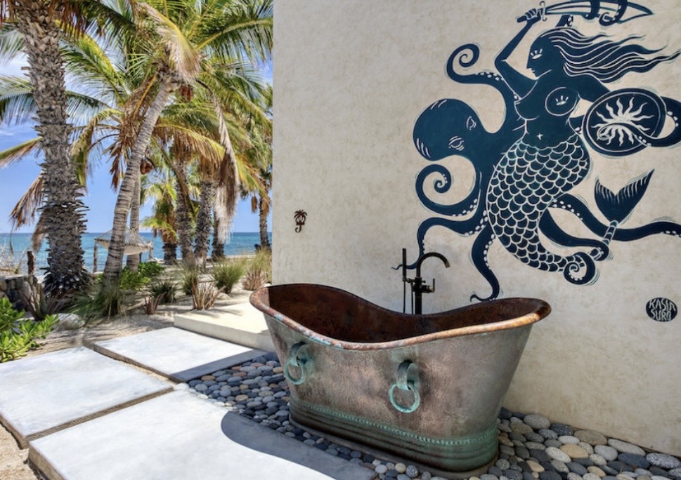 Copper wellness tub outdoors beside a hand-painted mermaid mural with ocean and palms beyond